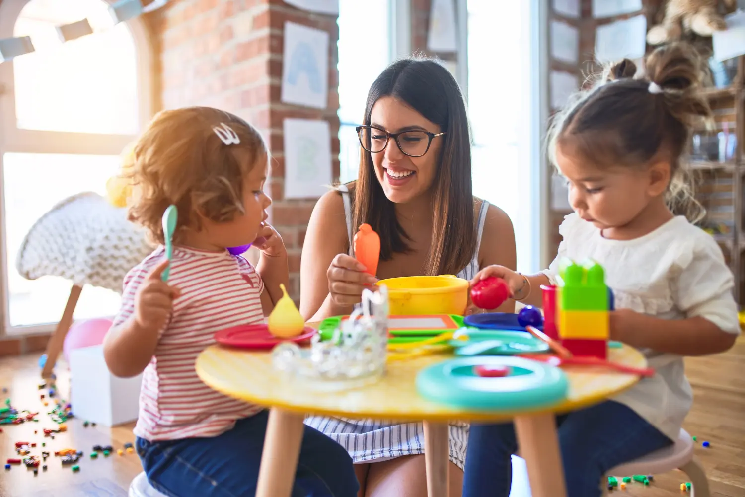 Junge schöne Lehrerin und Kleinkinder spielen im Kindergarten Mahlzeiten mit Plastiklebensmitteln und Besteckspielzeug